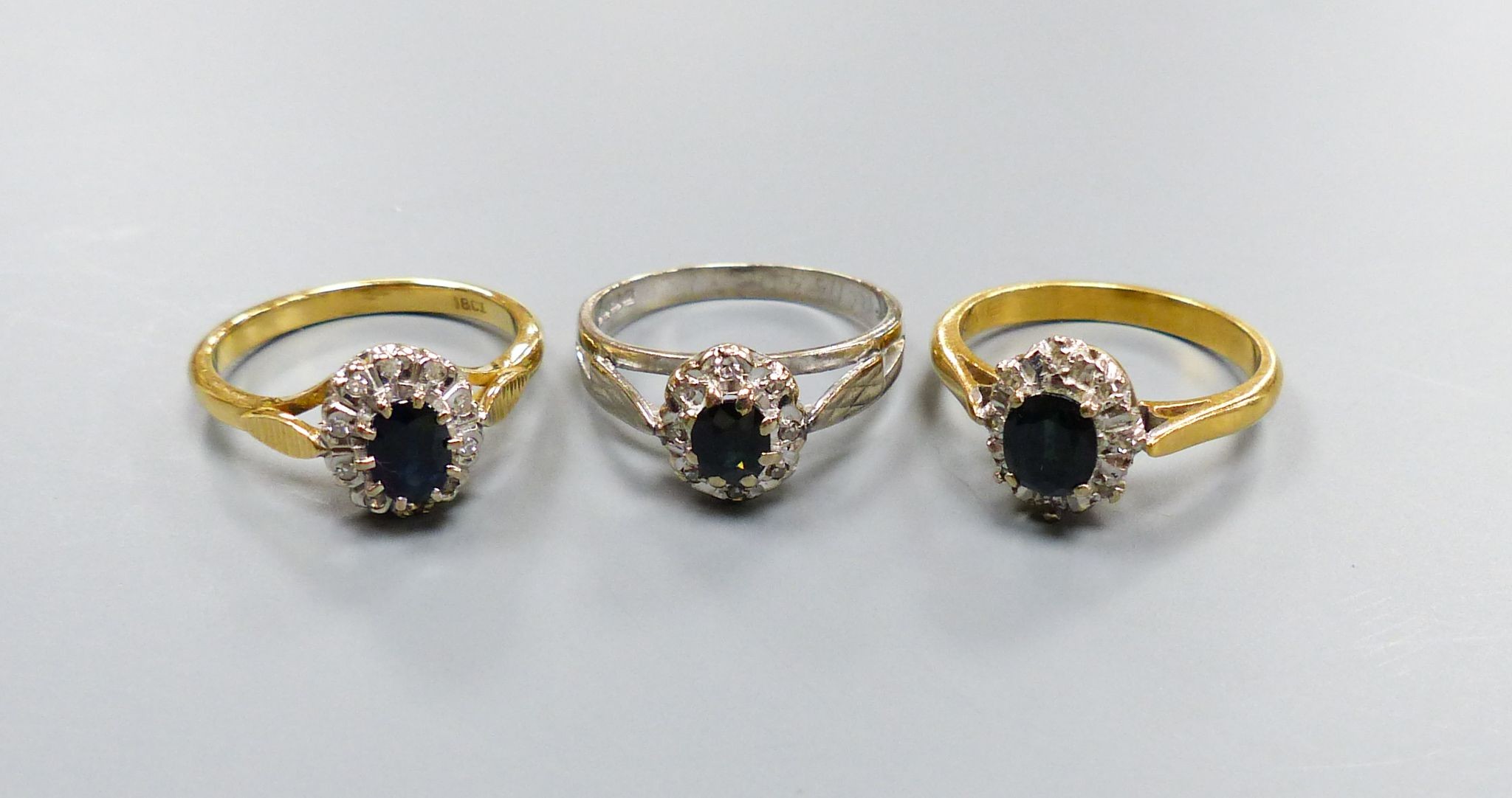 Three modern sapphire and diamond set oval cluster rings, including 18ct white gold, size M and 18ct, size L/M, (gross 7.2 grams and one yellow metal, size L/M, gross 3.8 grams.